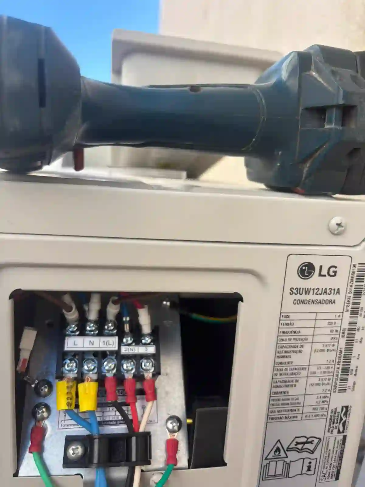 Detalhe da instalação elétrica de uma condensadora, com bornes, fiação e parafusadeira apoiada sobre o equipamento.